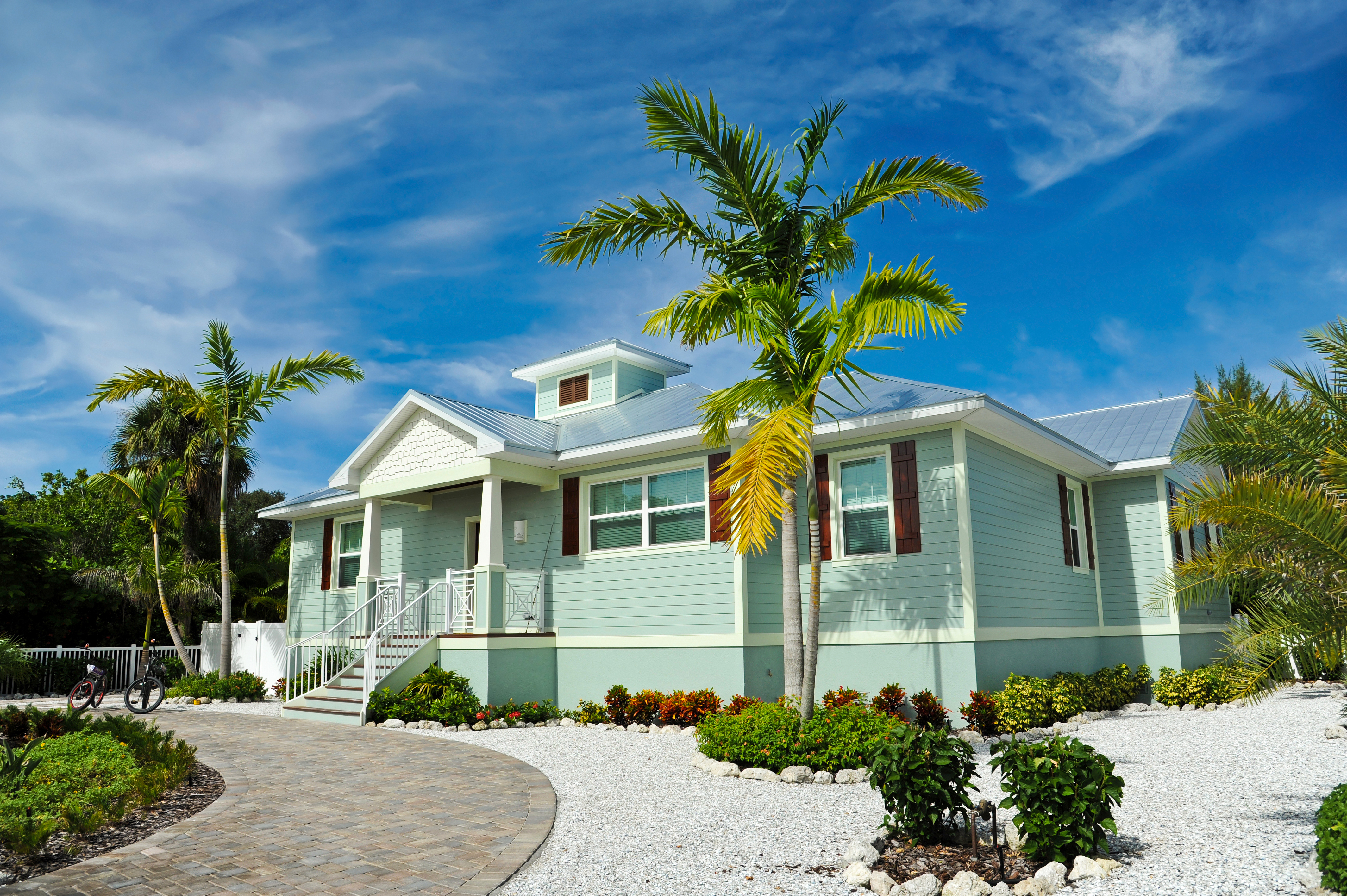 front view of a sea green beachhouse with palm trees and pebbles lining the stone driveway